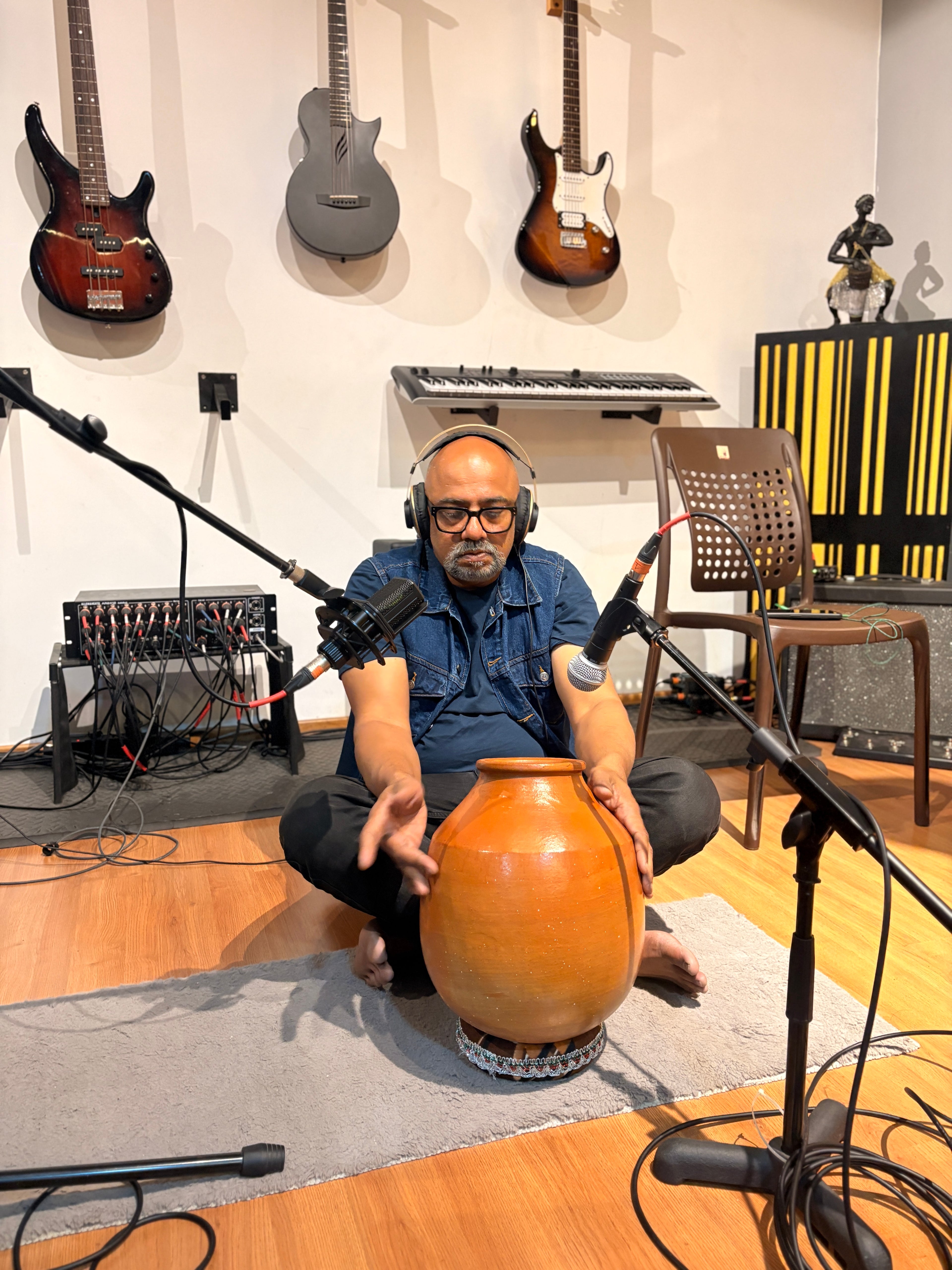 Man playing a large wooden drum in a music studio with guitars and equipment in the background.