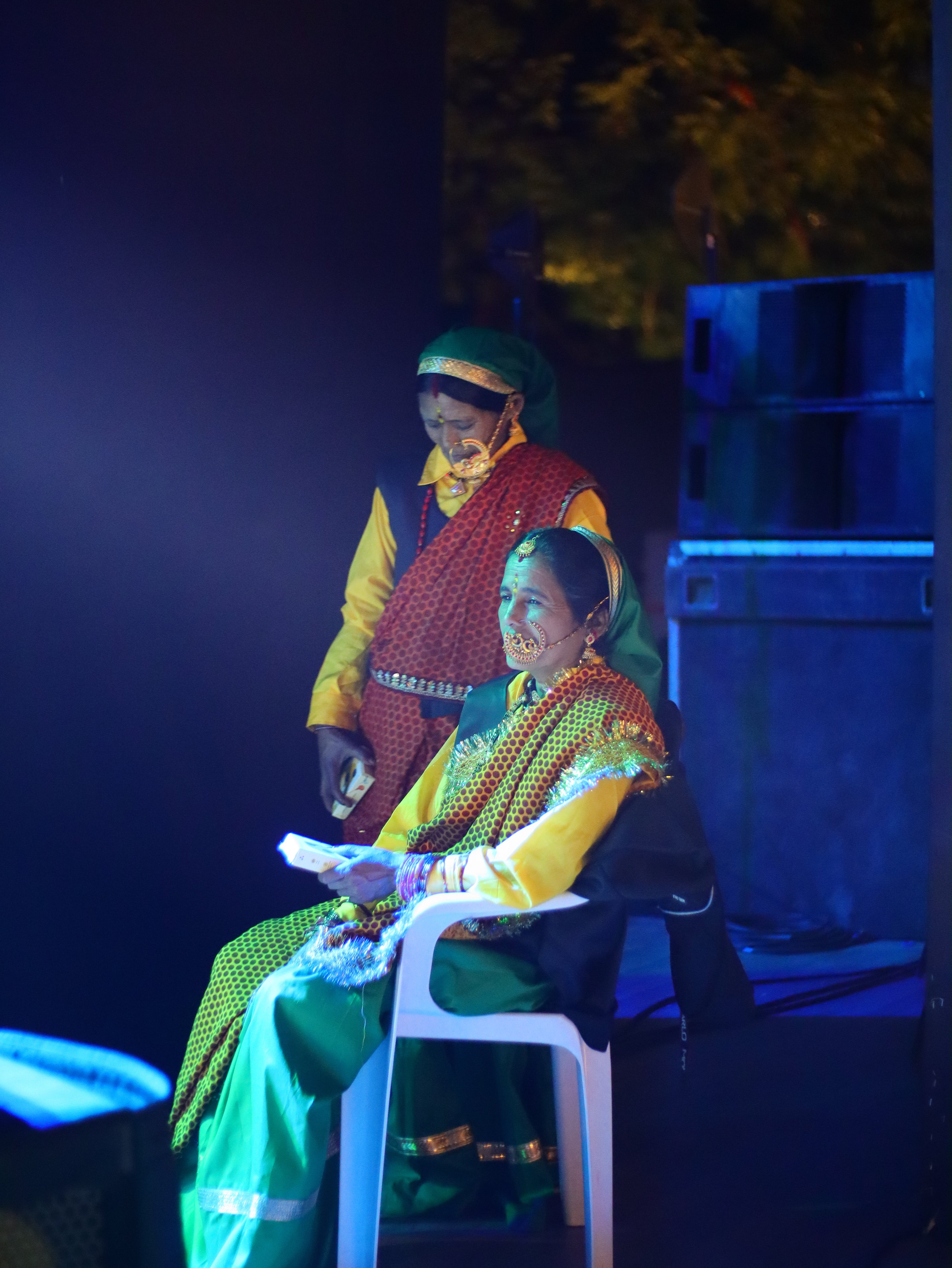 A person in a vibrant green, yellow, and red patterned outfit with a matching headwrap, seated on a white chair in a dimly lit stage-like environment with blue lighting.